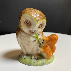 May include: A ceramic figurine of an owl and a squirrel. The owl has brown and cream-colored feathers and large eyes. The squirrel is orange with a bushy tail. Both are on a green base, holding a sprig of leaves.
