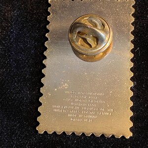 Pu&ograve; includere: Una spilla dorata a forma di francobollo. La spilla ha una superficie strutturata e una chiusura sul retro. Il testo sulla spilla recita "WINCO INTERNATIONAL" e "MADE IN TAIWAN".