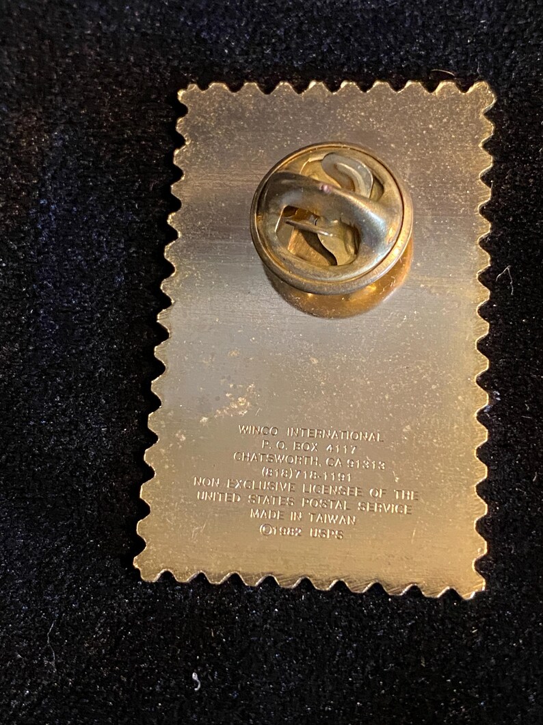 Pu&ograve; includere: Una spilla dorata a forma di francobollo. La spilla ha una superficie strutturata e una chiusura sul retro. Il testo sulla spilla recita "WINCO INTERNATIONAL" e "MADE IN TAIWAN".