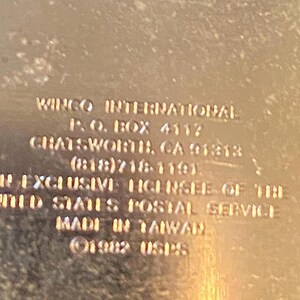 Pu&ograve; includere: Un oggetto metallico color oro con un bordo smerlato, con testo in rilievo. Il testo include le parole "WINGO INTERNATIONAL", "MADE IN TAIWAN" e "1982 USPS". L'articolo &egrave; un prodotto con licenza del United States Postal Service.