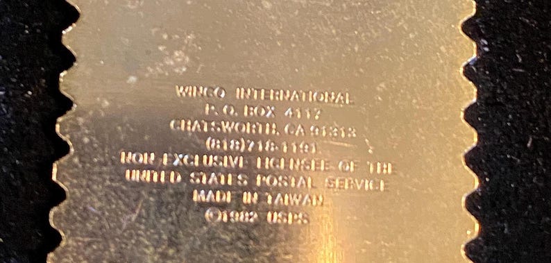 Pu&ograve; includere: Un oggetto metallico color oro con un bordo smerlato, con testo in rilievo. Il testo include le parole "WINGO INTERNATIONAL", "MADE IN TAIWAN" e "1982 USPS". L'articolo &egrave; un prodotto con licenza del United States Postal Service.