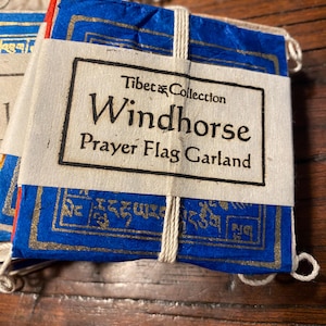 May include: A stack of blue and gold Tibetan Windhorse Prayer Flag Garlands. The flags are tied together with a white string and feature gold lettering and designs. The label reads "Tibet Collection Windhorse Prayer Flag Garland."