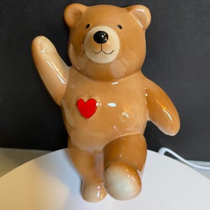 May include: A ceramic teddy bear figurine in shades of brown with a red heart on its chest. The bear is posed with one arm raised in a waving gesture and the other leg extended. The bear has a glossy finish and a smiling face.