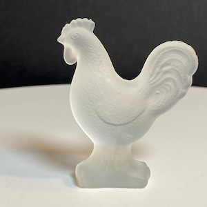 May include: A frosted glass rooster figurine. The sculpture features a detailed comb, wattle, and tail feathers. The rooster stands on a rectangular base. The figurine is a decorative item.