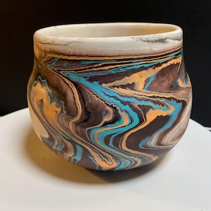 May include: A rounded ceramic bowl featuring a marbled pattern of swirling dark brown, bright turquoise, and muted orange colors. The vessel has a wide, open mouth with a smooth, light-colored rim that transitions into the intricate, fluid design.