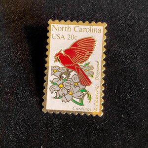 Pu&ograve; includere: Spilla rettangolare a forma di francobollo, raffigurante un uccello cardinale rosso con le ali spiegate, posato su fiori di corniolo bianco. Il testo "North Carolina USA 20c" &egrave; stampato in alto, con "Cardinal" in basso.