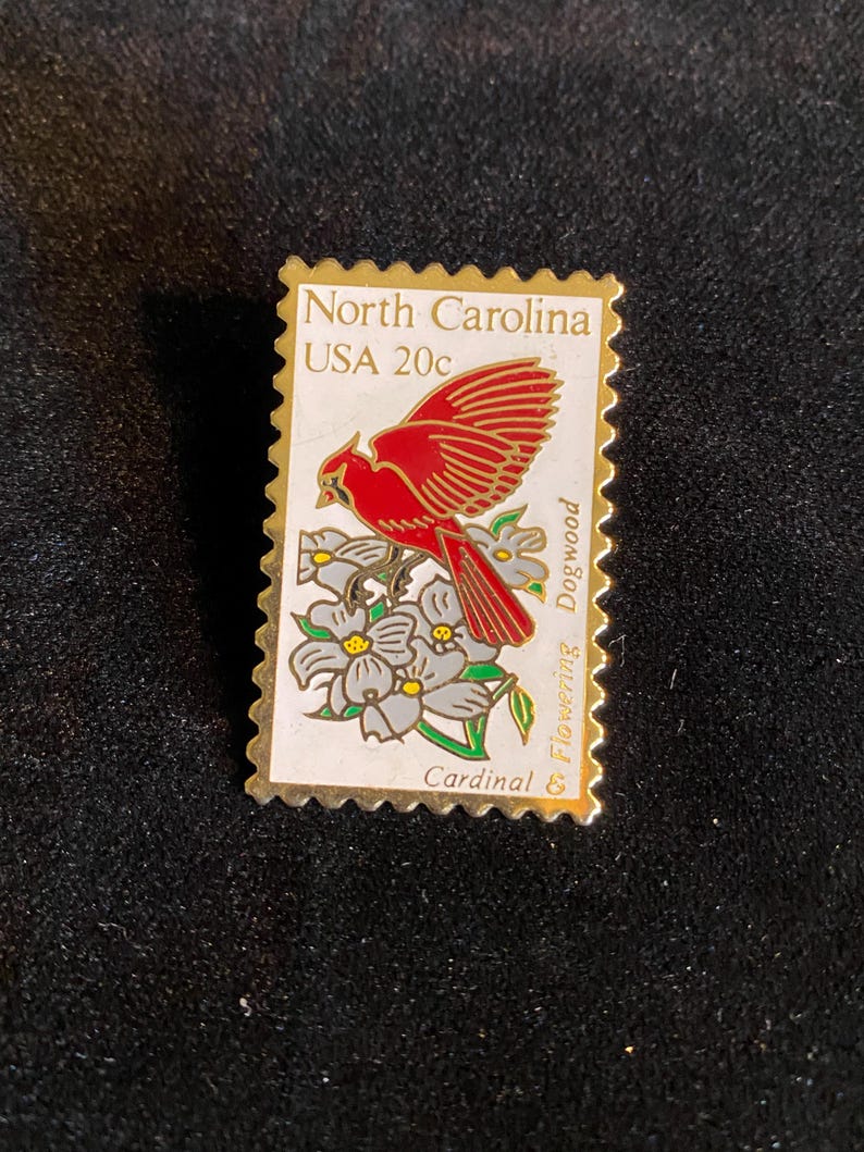Pu&ograve; includere: Spilla rettangolare a forma di francobollo, raffigurante un uccello cardinale rosso con le ali spiegate, posato su fiori di corniolo bianco. Il testo "North Carolina USA 20c" &egrave; stampato in alto, con "Cardinal" in basso.
