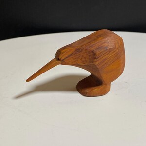 May include: A small, hand-carved wooden figurine of a kiwi bird. The sculpture features a smooth, light brown wood grain with a long, pointed beak and a rounded body, capturing the distinct silhouette of the flightless bird in a solid form.