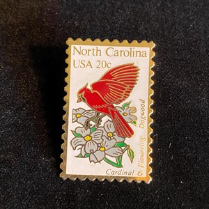 Pu&ograve; includere: Spilla con bordi dorati a forma di francobollo North Carolina USA 20c. Il francobollo raffigura un cardinale rosso appollaiato su rami di corniolo in fiore con fiori grigi e bianchi.