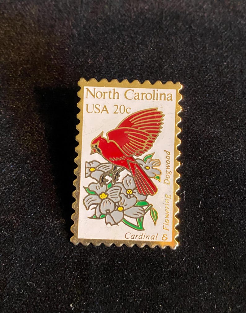 Pu&ograve; includere: Spilla con bordi dorati a forma di francobollo North Carolina USA 20c. Il francobollo raffigura un cardinale rosso appollaiato su rami di corniolo in fiore con fiori grigi e bianchi.