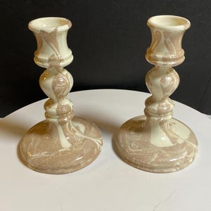 May include: A pair of matching candlestick holders featuring a glossy, cream and tan marbled pattern. Each holder has a wide circular base, a slender stem with decorative rounded nodes, and a flared top opening designed to hold a single taper candle.