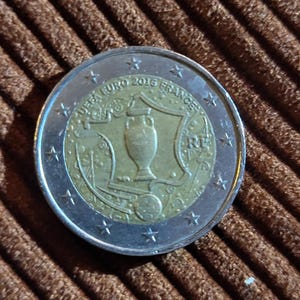 Könnte beinhalten: Eine silberne und goldfarbene Euro-Münze mit einem Design, das an das UEFA-Europameisterschaftsturnier 2016 in Frankreich erinnert. Die Münze zeigt eine Trophäe und den Text "UEFA EURO 2016 FRANCE".