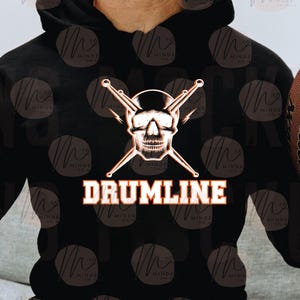 Puede incluir: Sudadera con capucha negra con un diseño de calavera, baquetas cruzadas y la palabra "DRUMLINE" en naranja y blanco. El diseño está centrado en la parte delantera. La sudadera tiene una capucha con cordón y un bolsillo canguro.