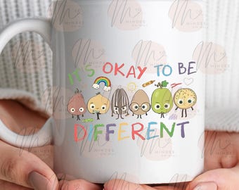 May include: White ceramic mug featuring the text "It's Okay to Be Different" in colorful letters. The mug showcases cartoon illustrations of various food items with faces, including a rainbow, and a crayon. The mug is held by a person.