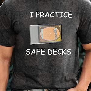 May include: Dark gray t-shirt with the text "I PRACTICE SAFE DECKS" in white. A card is visible in a clear plastic sleeve. The card has a gold and brown design with the word "Magic" visible.