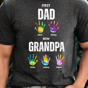 Puede incluir: Camiseta gris oscuro con el texto "FIRST DAD NOW GRANDPA". La camiseta presenta huellas de manos coloridas con los nombres: Matt, Ashley, Jaden, Juliana y Jaycee. Un regalo personalizado.