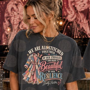 Op de afbeelding: Een antracietgrijs t-shirt met een afbeelding van een patchwork-jas, een vlinder en de tekst: WE ARE ALL STITCHED TOGETHER BY OUR STRUGGLES AND MADE BEAUTIFUL BY OUR RESILIENCE Dolly Parton. Het ontwerp bevat sterren en hartjes.