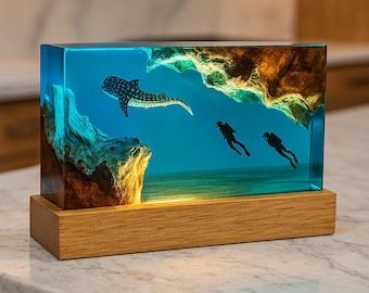 May include: A rectangular resin art piece depicting an underwater scene with a whale shark and two scuba divers. The resin is a clear blue, with a wooden base. The scene includes rock formations and a sandy ocean floor.