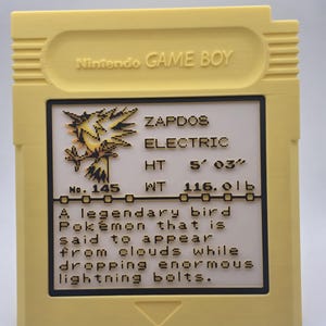 May include: A yellow Nintendo Game Boy cartridge featuring a Zapdos Pok&eacute;mon card design. The card includes the text "ZAPDOS ELECTRIC" and details about the Pok&eacute;mon's height and weight, 5' 03" and 116.0 lb, respectively. The cartridge has a black border.