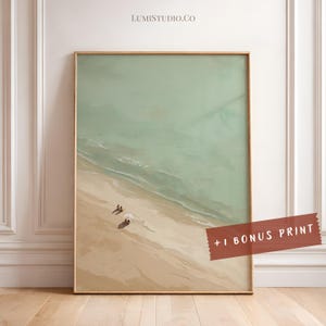 May include: A vertical rectangular wall art print featuring an aerial view of a sandy beach meeting a pale green ocean. Two small figures and a white umbrella are visible on the sand. A red banner in the corner displays the text + 1 BONUS PRINT.