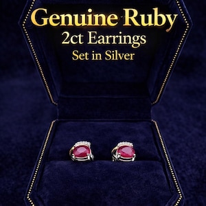 May include: A pair of genuine ruby earrings set in silver, displayed in a dark blue velvet jewelry box. The earrings feature a large, oval-cut ruby gemstone with a silver setting. The box has gold trim and the text "Genuine Ruby 2ct Earrings Set in Silver".