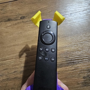 May include: A black remote control with buttons for navigation, volume, and playback. The remote is held in a purple 3D printed stand with yellow accents.