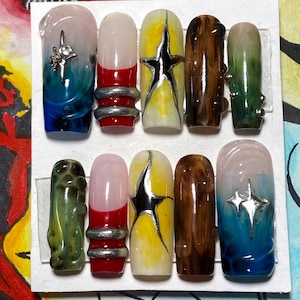May include: Set of ten false nails with different designs. The nails are painted with various colors and patterns, including red, yellow, green, blue, and brown. Some nails have silver accents and rhinestones.