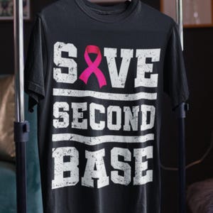 May include: Black t-shirt with the words "SAVE SECOND BASE" in distressed white lettering. A pink ribbon is featured above the word "SECOND". The shirt is a crew neck style.