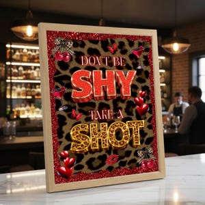 以下が含まれることがあります： 「DON'T BE SHY TAKE A SHOT」の文字が赤と金色の文字で書かれた額入りのサイン。背景はヒョウ柄模様。赤いハート、蝶、さくらんぼ、カクテルグラスなどの装飾要素があります。フレームは薄茶色です。