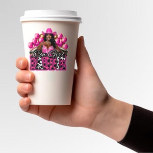 K&ouml;nnte beinhalten: Ein wei&szlig;er Pappbecher mit einer Grafik einer Frau in einem rosa Cowboyhut und -outfit, mit dem Text "Cow Girl Baddie" in Rosa und Schwarz. Rosa Ballons sind im Hintergrund.