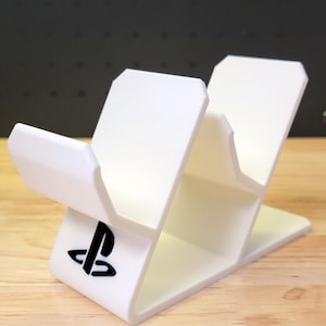 May include: White plastic stand with two slots for holding video game controllers. The stand has a PlayStation logo on the front.
