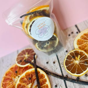 May include: A clear plastic pouch containing dried orange slices, whole peppercorns, and a vanilla bean. A circular white label on the front features the text "Morgan's Melts" and "Wildly Spring" with a floral graphic. Loose dried citrus and spices surround the bag.