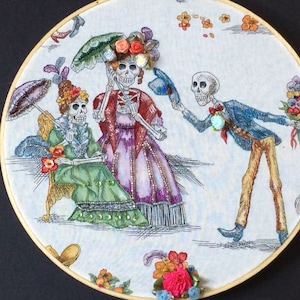 May include: An embroidered artwork in a wooden hoop depicts a Day of the Dead scene. Three skeletons are dressed in colorful attire, with one offering flowers. The background is a patterned fabric with floral and shoe motifs.