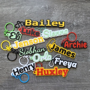 May include: A collection of colorful custom name keychains featuring the names Bailey, Luke, Sienna, Eli, Jenson, Archie, Siobhan, James, Orla, Freya, Henry, and Huxley. Each keychain is made of layered plastic with attached metal rings and chains.