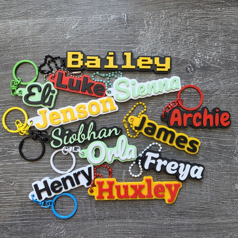 May include: A collection of colorful custom name keychains featuring the names Bailey, Luke, Sienna, Eli, Jenson, Archie, Siobhan, James, Orla, Freya, Henry, and Huxley. Each keychain is made of layered plastic with attached metal rings and chains.