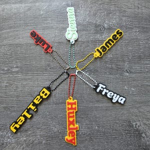May include: Six custom silicone name keychains arranged in a star pattern. The tags feature the names Sienna, Luke, James, Bailey, Huxley, and Freya in various colors including red, yellow, black, white, and light green, each with a matching ball chain.