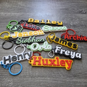 May include: A collection of custom 3D-printed name keychains in various colors including yellow, black, red, white, and light green. Each keychain features a raised name such as Bailey, Eli, Luke, Sienna, Jenson, Siobhan, James, Archie, Orla, Freya, Henry, and Huxley.