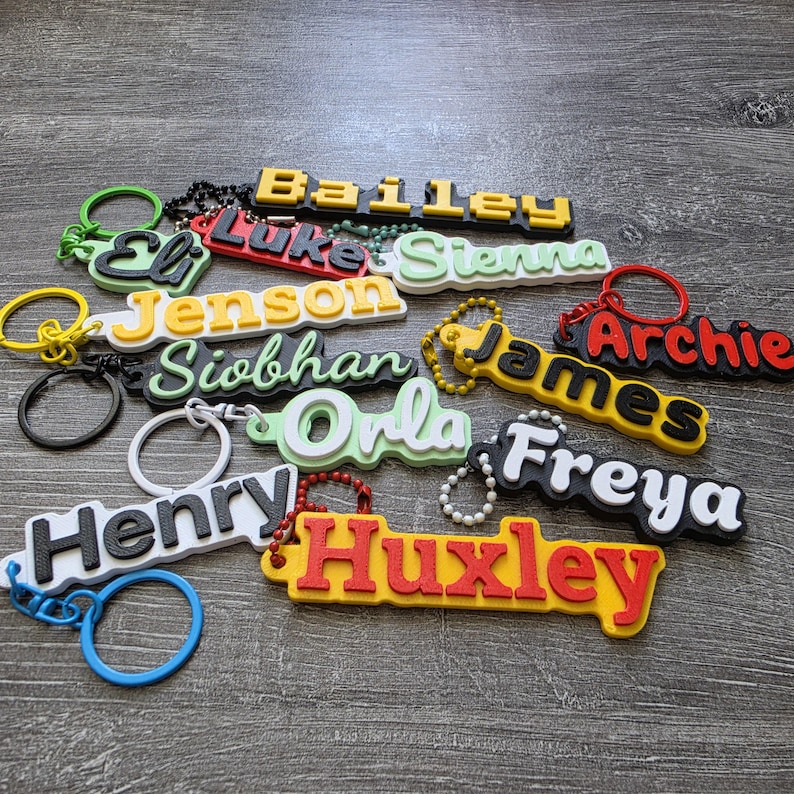 May include: A collection of custom 3D-printed name keychains in various colors including yellow, black, red, white, and light green. Each keychain features a raised name such as Bailey, Eli, Luke, Sienna, Jenson, Siobhan, James, Archie, Orla, Freya, Henry, and Huxley.
