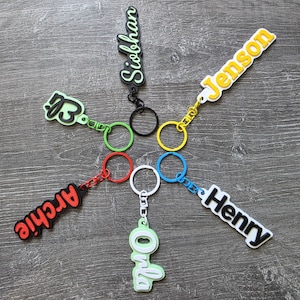 May include: Six personalized keychains arranged in a circle, each featuring a name in a raised, colorful font. The names include Siobhan, Jenson, Henry, Onla, Archie, and a script name, attached to matching colored metal split rings.