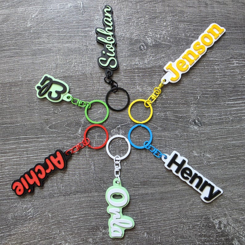 May include: Six personalized keychains arranged in a circle, each featuring a name in a raised, colorful font. The names include Siobhan, Jenson, Henry, Onla, Archie, and a script name, attached to matching colored metal split rings.