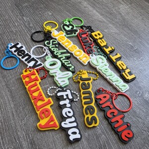 May include: A collection of colorful silicone keychains featuring raised lettering in various fonts and colors, including names such as Henry, Jenson, Siobhan, Luke, Bailey, Sienna, Huxley, Freya, James, and Archie, each attached to a metal ring.