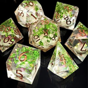 May include: A set of seven polyhedral dice. Each die is clear with green plant inclusions and copper-coloured numbers. The dice are arranged on a black surface, showcasing their unique design and intricate details. Suitable for tabletop games.