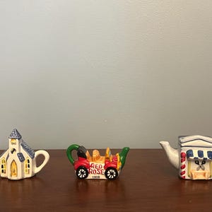 May include: Three miniature ceramic teapots. One is shaped like a church with a blue roof and yellow door. Another is shaped like a red car with the words "Red Rose 1908" on the side. The third is shaped like a white building with a blue awning and a striped red and white pole.