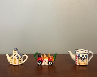 May include: Three miniature ceramic teapots. One is shaped like a church with a blue roof and yellow door. Another is shaped like a red car with the words "Red Rose 1908" on the side. The third is shaped like a white building with a blue awning and a striped red and white pole.