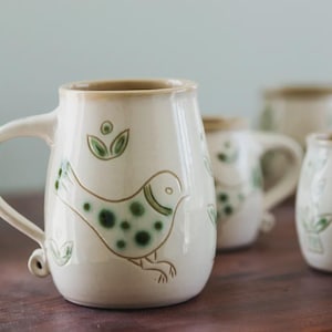 May include: Cream-colored ceramic mugs with a bird and leaf design in green and brown. The mugs have handles and are arranged on a wooden surface. The bird has a dotted pattern.