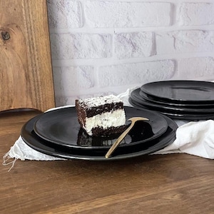 May include: A stack of black ceramic plates with a piece of chocolate cake with white frosting and a gold-colored serving utensil. The cake is topped with white sprinkles. A wooden cutting board is in the background.