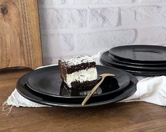 May include: A stack of black ceramic plates with a piece of chocolate cake with white frosting and a gold-colored serving utensil. The cake is topped with white sprinkles. A wooden cutting board is in the background.