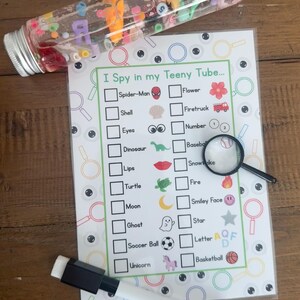 May include: A clear plastic tube filled with colorful beads, letters, and shapes, next to an "I Spy" game sheet with a magnifying glass and a dry-erase marker. The game sheet has a list of items to find.