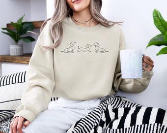 May include: A beige sweatshirt with a graphic of three dachshund dogs in a line. The dogs are outlined in black and are facing different directions.