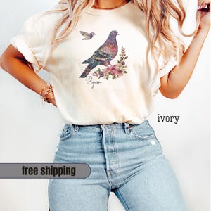May include: Ivory-colored t-shirt featuring a detailed illustration of a pigeon with floral accents and the word "Pigeon" in script. The design includes a smaller bird in flight. The shirt is paired with light blue jeans.